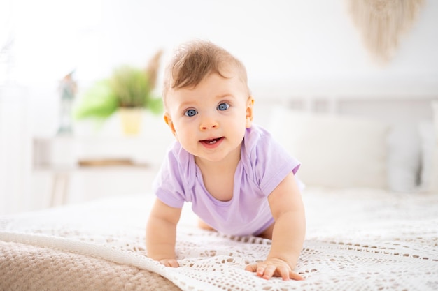 Un lindo bebé sano con un traje lila se arrastra sobre una cama con sábanas blancas en el dormitorio de la casa un retrato de un niño mira la cámara