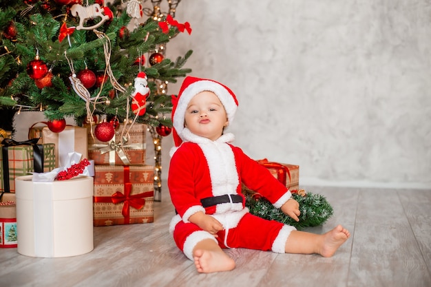Lindo bebé Santa se sienta en casa cerca del árbol de Navidad con regalos