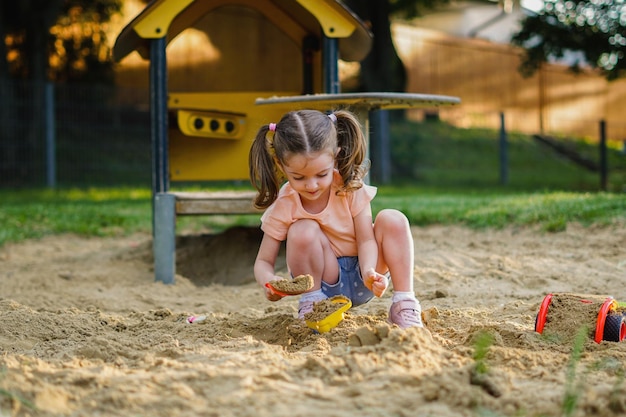 Lindo bebê se divertindo no dia ensolarado e quente de verão Menina bonita brincando na areia no playground ao ar livre