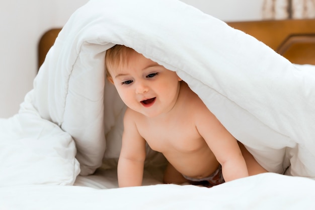 Lindo bebé sentado en una cama envuelto en una manta blanca. el bebé tiene 11 meses.