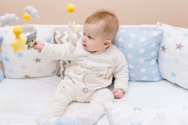 Lindo bebé sentado en una cama redonda blanca. Guardería ligera para niños pequeños. Juguetes para cuna infantil. Niño sonriente que juega con el móvil del fieltro en dormitorio soleado.