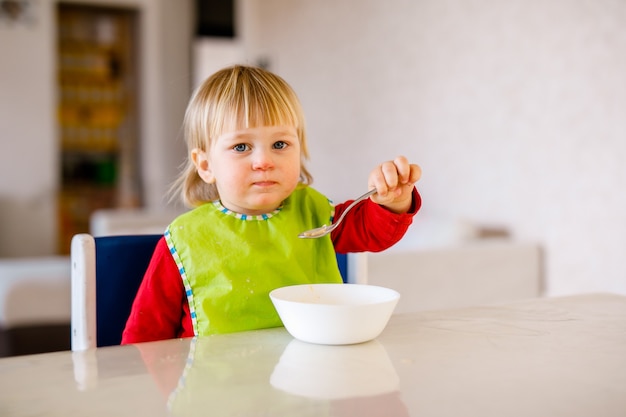 Lindo bebé sentado en la silla alta para niños y comiendo vegetales solo en la cocina blanca.