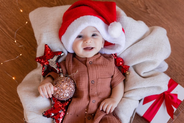 Un lindo bebé con un sombrero de Papá Noel está tirado en una manta en el suelo y sosteniendo una bola de Navidad en la mano. felices vacaciones. invierno, año nuevo. foto de alta calidad