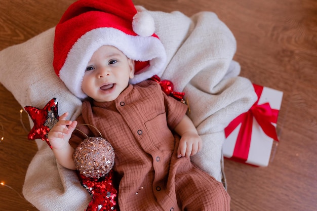 Un lindo bebé con un sombrero de Papá Noel está tirado en una manta en el suelo y sosteniendo una bola de Navidad en la mano. felices vacaciones. invierno, año nuevo. foto de alta calidad