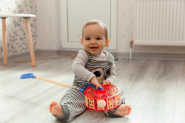 Foto lindo bebé sonriente de 12 meses se sienta en el suelo de su habitación y juega con juguetes.