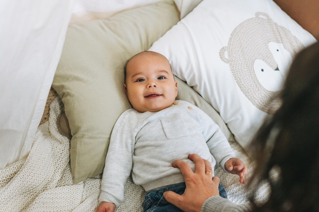 Lindo bebé sonriente en beige de 56 meses mirando a la cámara con la madre en la cama con manta tejida