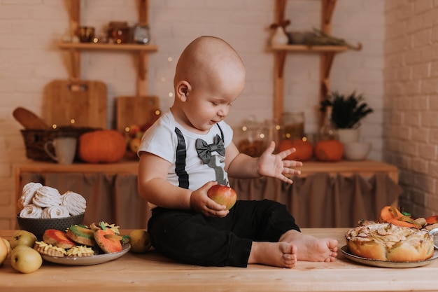 Lindo bebé sonriente sentado en una mesa de cocina de madera sosteniendo una manzana en sus manos. primer cumpleaños en un ambiente festivo. merienda saludable para niños. tortas caseras, golosinas para invitados. foto de alta calidad