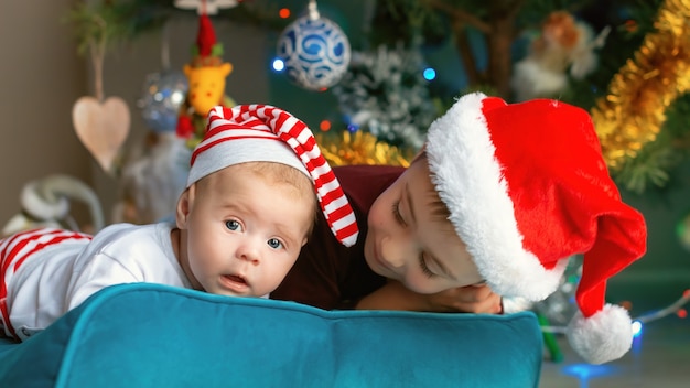 Lindo bebé con su hermano mayor en el contexto de un árbol de Navidad. Niño disfrazado de elfo blanco rojo con su hermano con sombrero de santa jugando juntos.