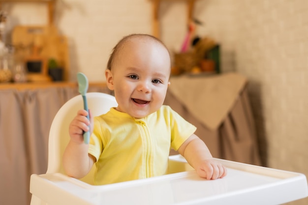 Un lindo bebé con un traje amarillo está sentado en una silla alta en la cocina de la casa con un