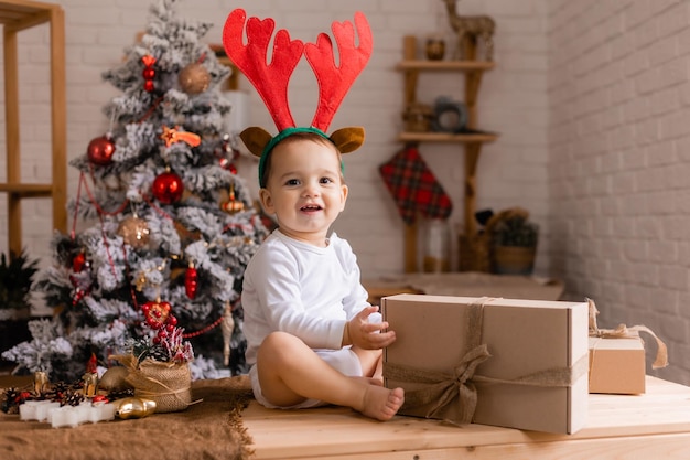 Un lindo bebé con un traje blanco y cuernos de reno está sentado con regalos en la cocina en casa para Navidad, el niño mira los regalos para el nuevo año
