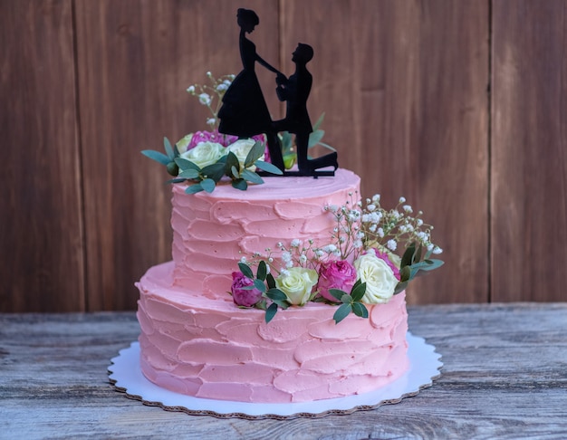 Lindo bolo de casamento de duas camadas com creme de queijo rosa, decorado com rosas vivas e a figura de um casal apaixonado, em uma mesa de madeira