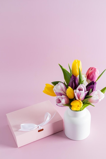 Lindo buquê de tulipas coloridas em um vaso de cerâmica branco em um fundo rosa com uma caixa de presente festiva vista vertical um espaço de cópia