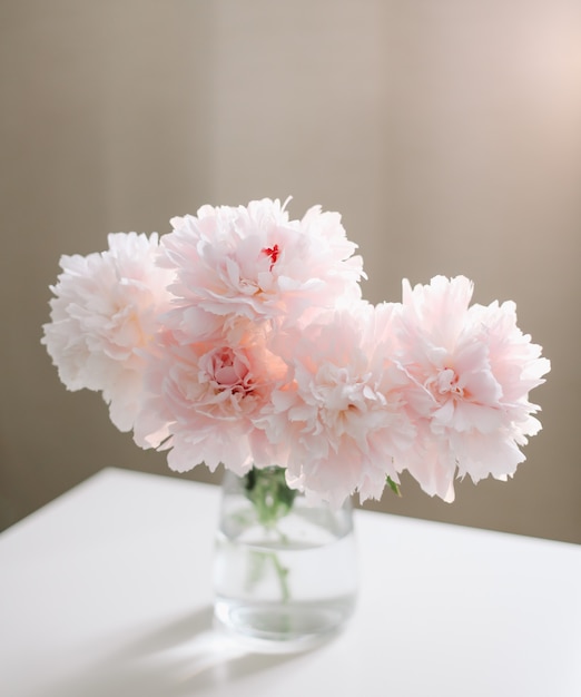 Lindo buquê rosa claro de peônias rosa em um vaso de vidro na mesa branca perto de uma janela