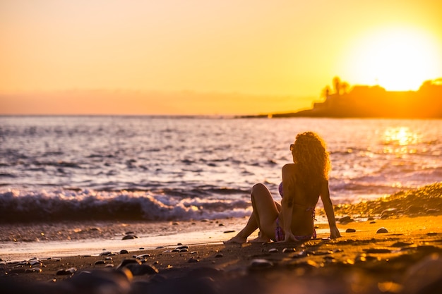 Lindo cabelo encaracolado modelo legal mulher de meia-idade jovem sentado na praia, apreciando a liberdade e o pôr do sol relaxado com cores douradas. diversão ao ar livre para senhora em férias