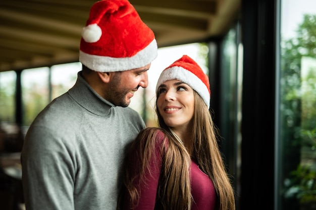 Lindo casal abraçando usando chapéus de papai noel perto de uma janela