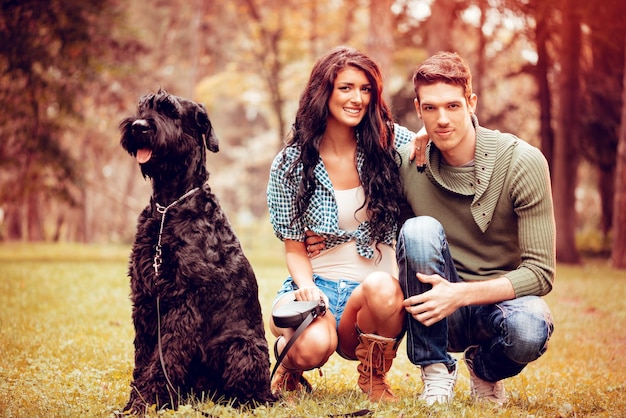 Lindo casal adorável com um cachorro, um schnauzer gigante preto, desfrutando no parque em cores de outono. Olhando para a câmera.