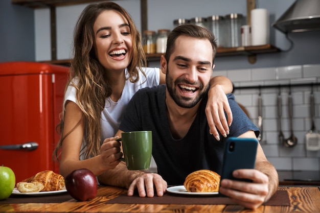 Lindo casal alegre tomando café da manhã na cozinha, usando telefone celular