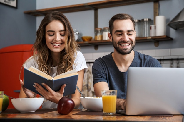 Lindo casal alegre tomando café da manhã na cozinha, usando um laptop, lendo um livro