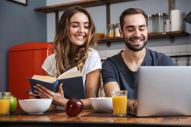 Lindo casal alegre tomando café da manhã na cozinha, usando um laptop, lendo um livro