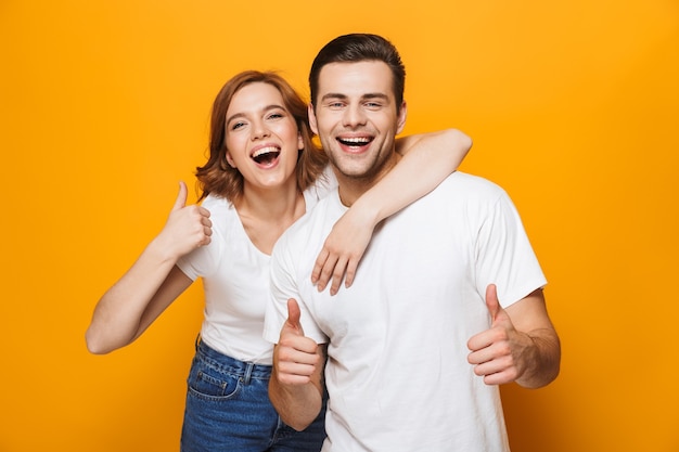 Lindo casal animado usando camisetas brancas, isolado na parede amarela, mostrando os polegares para cima