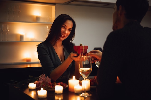 Lindo casal apaixonado jantando à luz de velas em casa, bebendo vinho, mulher recebendo uma caixa de presente