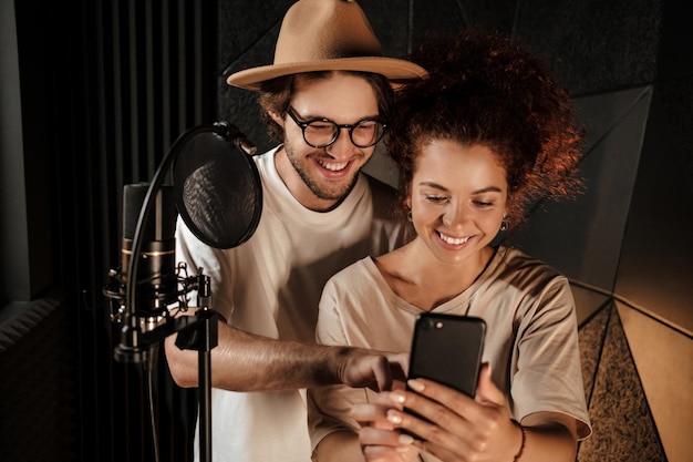 Lindo casal de cantores estilosos felizmente usando telefone trabalhando juntos no estúdio de gravação de som moderno