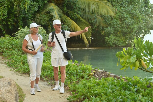 Lindo casal de idosos felizes descansando em um resort tropical com mochilas, homem apontando pela mão