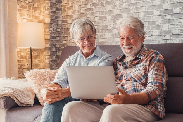 Lindo casal de idosos sentados no sofá usando laptop juntos comprando e navegando na rede Duas pessoas maduras na sala de estar curtindo tecnologia