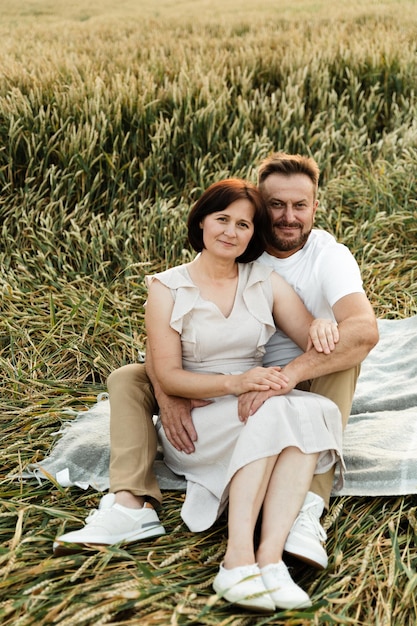 Lindo casal de meia-idade em um campo de trigo, sentado na colcha e se abraçando ternamente ao pôr do sol. pessoas mais velhas apaixonadas. amantes fazendo piquenique no campo