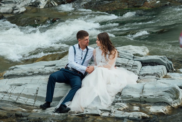 Lindo casal de noivos beijando e abraçando perto da margem de um rio de montanha com pedras