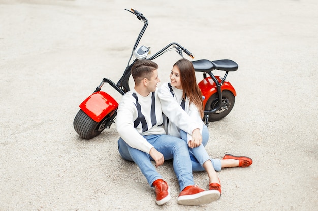 Foto lindo casal em roupas jeans da moda descansando perto de uma bicicleta elétrica vermelha na areia branca