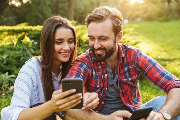 Lindo casal homem e mulher vestidos com roupas casuais, usando telefones celulares, enquanto descansam no parque verde