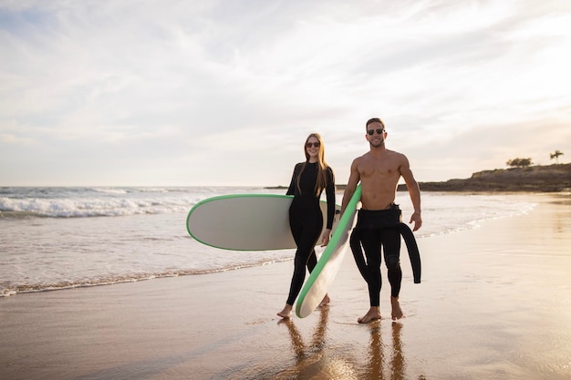 Lindo casal jovem caminhando na praia com pranchas de surf nas mãos no pôr do sol