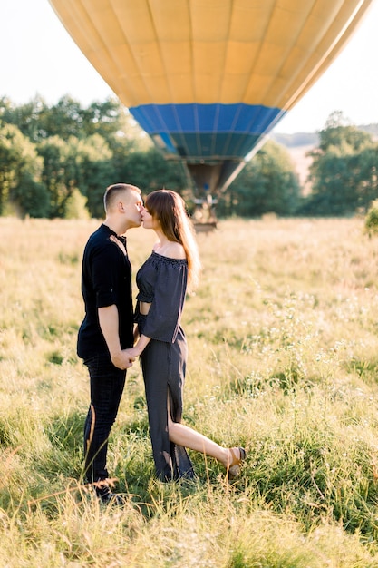 Lindo casal jovem caucasiano apaixonado, de pé no campo de verão, de mãos dadas e beijando. Balão de ar quente colorido ao nascer do sol