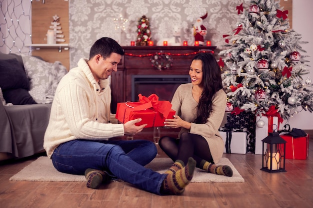 Lindo casal jovem com caixa de presente, sentado perto da árvore de Natal. Casa aconchegante. Casal feliz.