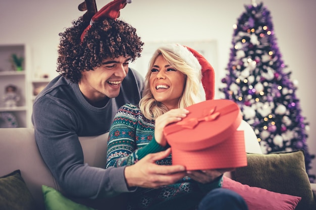 Lindo casal jovem comemorando em casa. Homem bonito está dando uma caixa de presente para sua namorada.