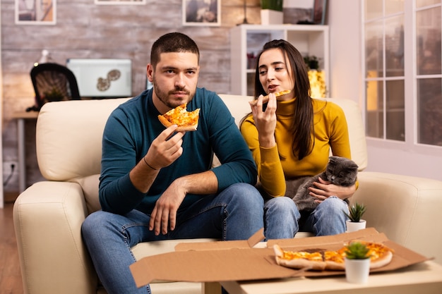 Lindo casal jovem comendo pizza enquanto assiste TV na sala de estar sentado no sofá