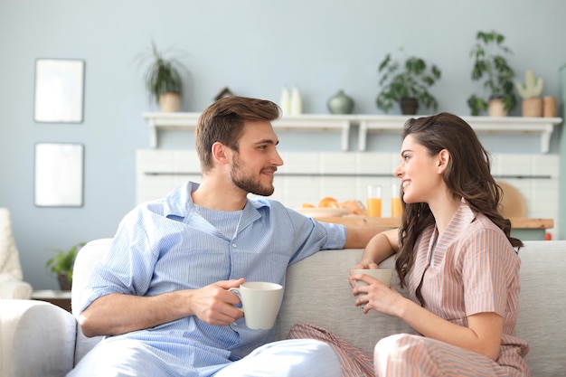 Lindo casal jovem de pijama está olhando um para o outro e sorrindo em um sofá na sala de estar.