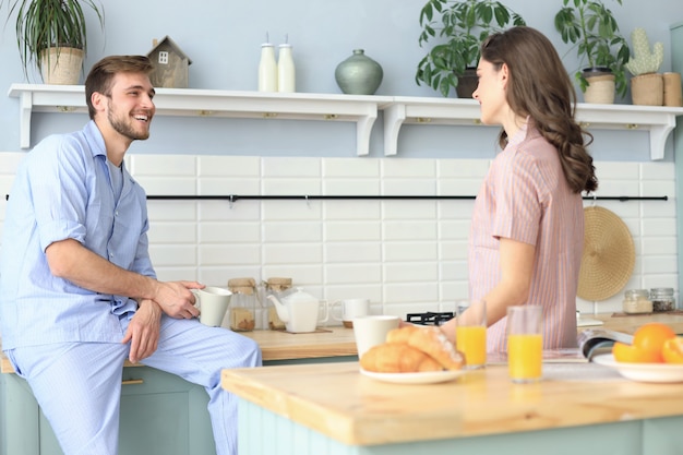 Lindo casal jovem de pijama está olhando um para o outro e sorrindo enquanto cozinha na cozinha em casa.