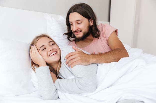 Lindo casal jovem deitado na cama em casa, sorrindo, acordando a namorada pela manhã