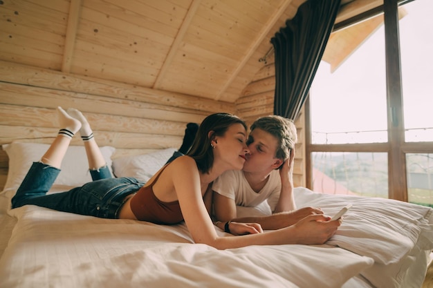 Lindo casal jovem deitado na cama em um quarto de hotel com um smartphone na mão