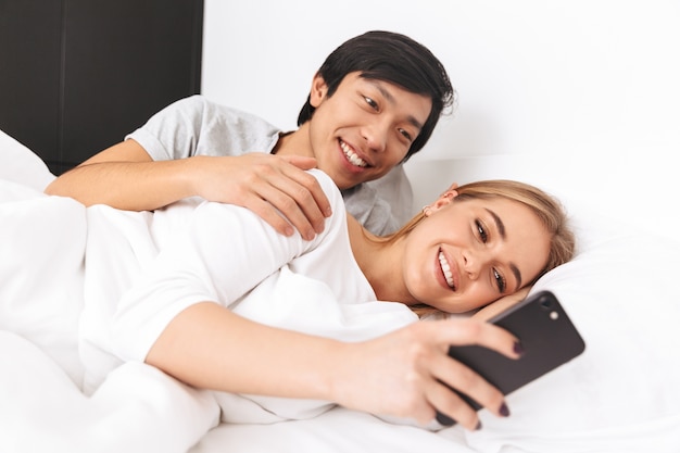 Lindo casal jovem, deitado na cama, jovem usando telefone celular