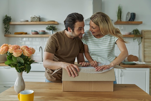 Lindo casal jovem desembalando a caixa e sorrindo em pé na cozinha doméstica