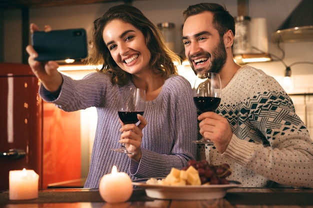 Lindo casal jovem e feliz passando uma noite romântica juntos em casa, bebendo vinho tinto, tirando uma selfie