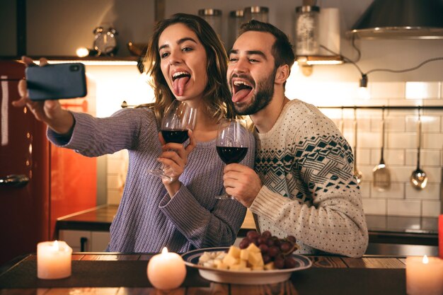 Lindo casal jovem e feliz passando uma noite romântica juntos em casa, bebendo vinho tinto, tirando uma selfie