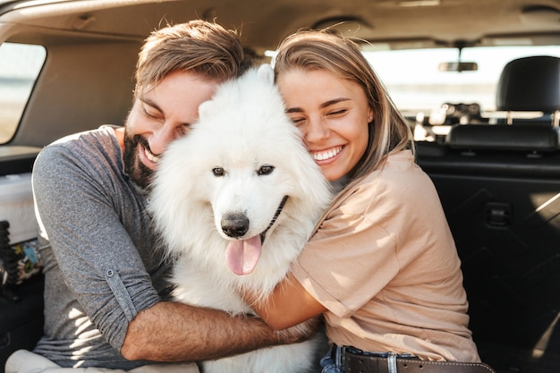 Lindo casal jovem e feliz sentado no banco de trás do carro na praia, brincando com o cachorro