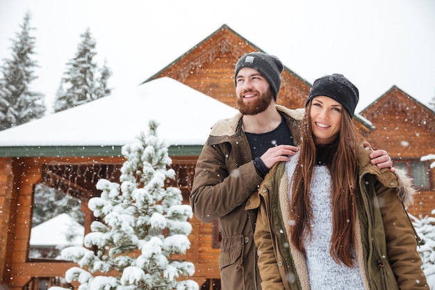 Lindo casal jovem em pé perto de uma cabana em tempo de neve