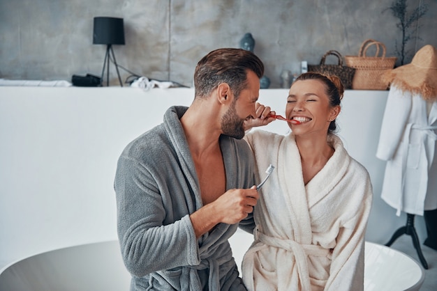 Foto lindo casal jovem em roupões de banho sorrindo e limpando os dentes enquanto faz a rotina matinal