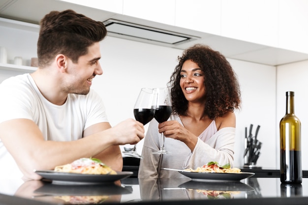 Lindo casal jovem multiétnico jantando romântico em casa, bebendo vinho tinto e comendo macarrão, brindando