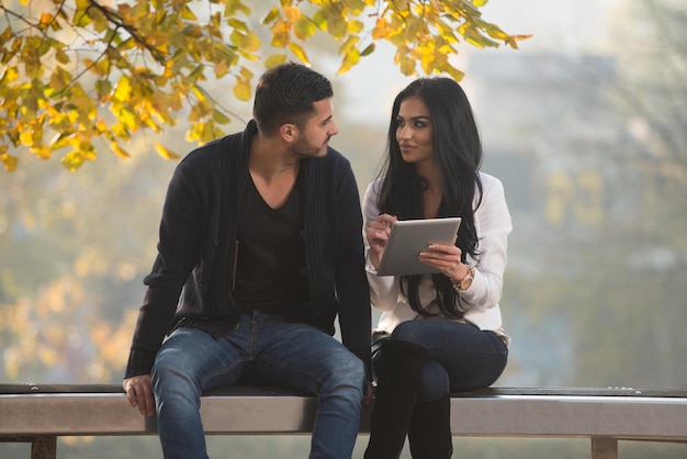 Lindo casal jovem sentado no banco no parque em um lindo dia de outono eles estão usando a Internet via tablet digital